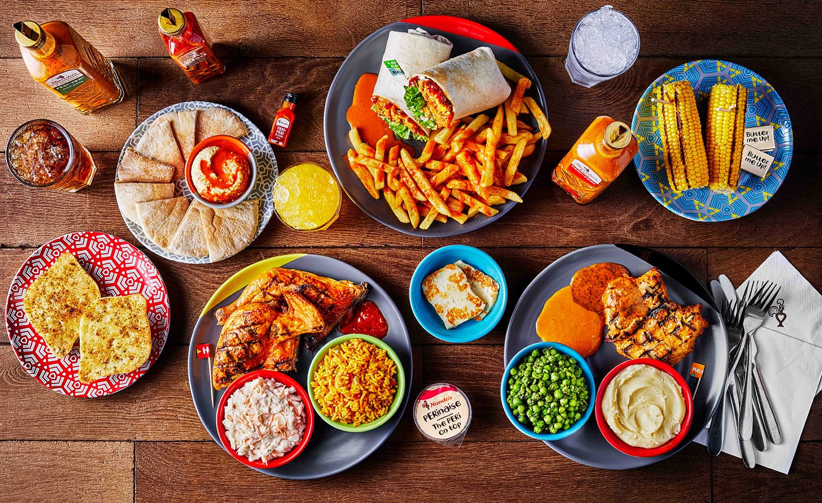 A colourful spread of Nando’s food, including grilled chicken, PERi-PERi fries, wraps, corn on the cob, garlic bread, hummus with pitta, sides, drinks, and their signature sauces.
