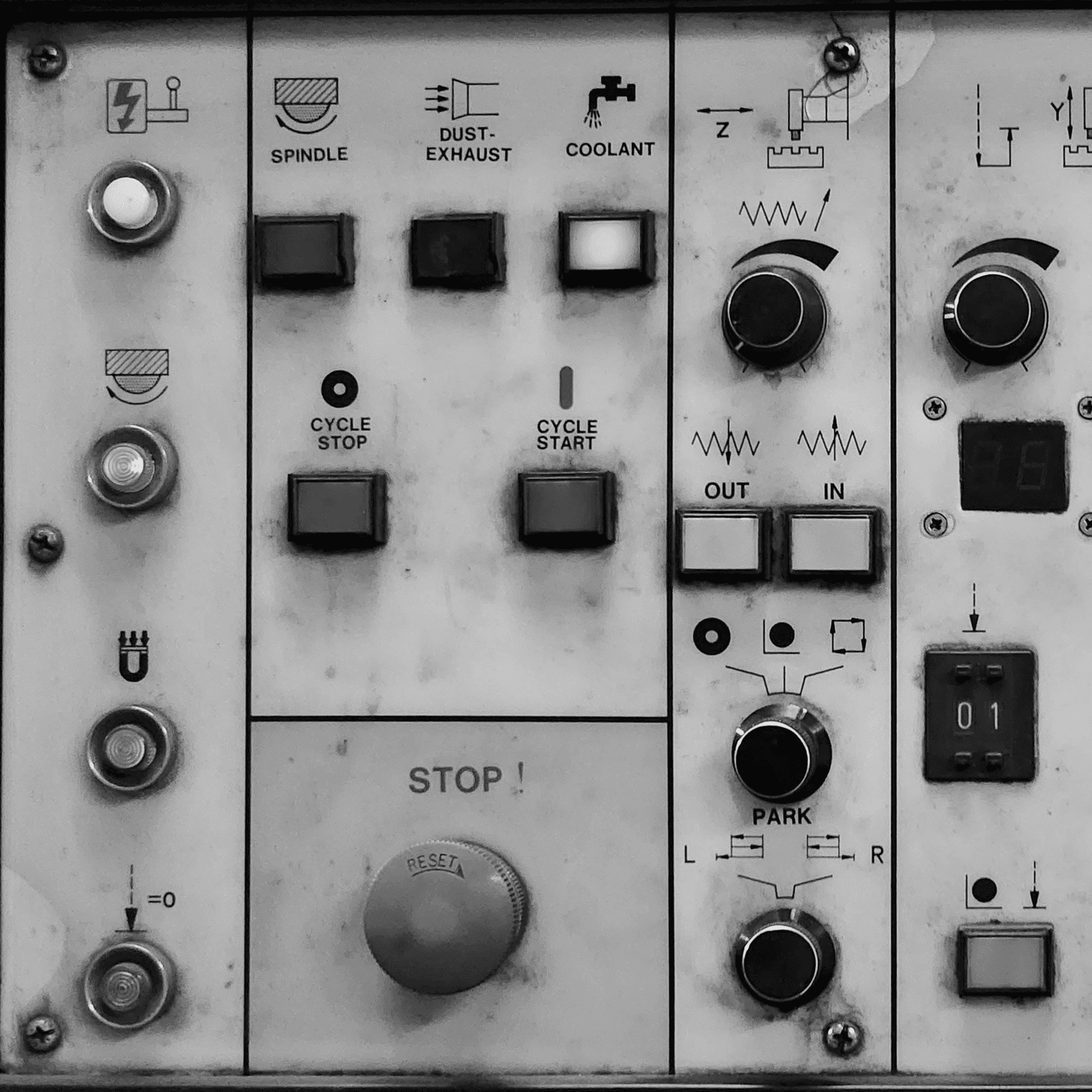 A Black-and-white close-up photograph of an industrial machine control panel, with labelled buttons, switches, dials, and a large stop button.