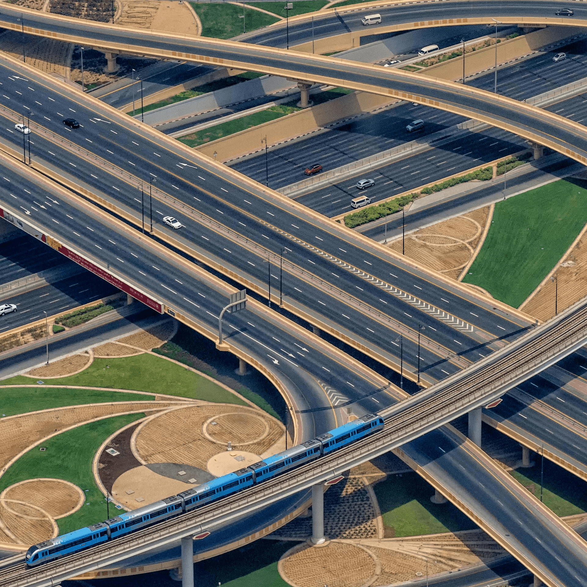 Aerial view photograph of a multilayer road interchange, with a blue metro train crossing the foreground on an elevated track.